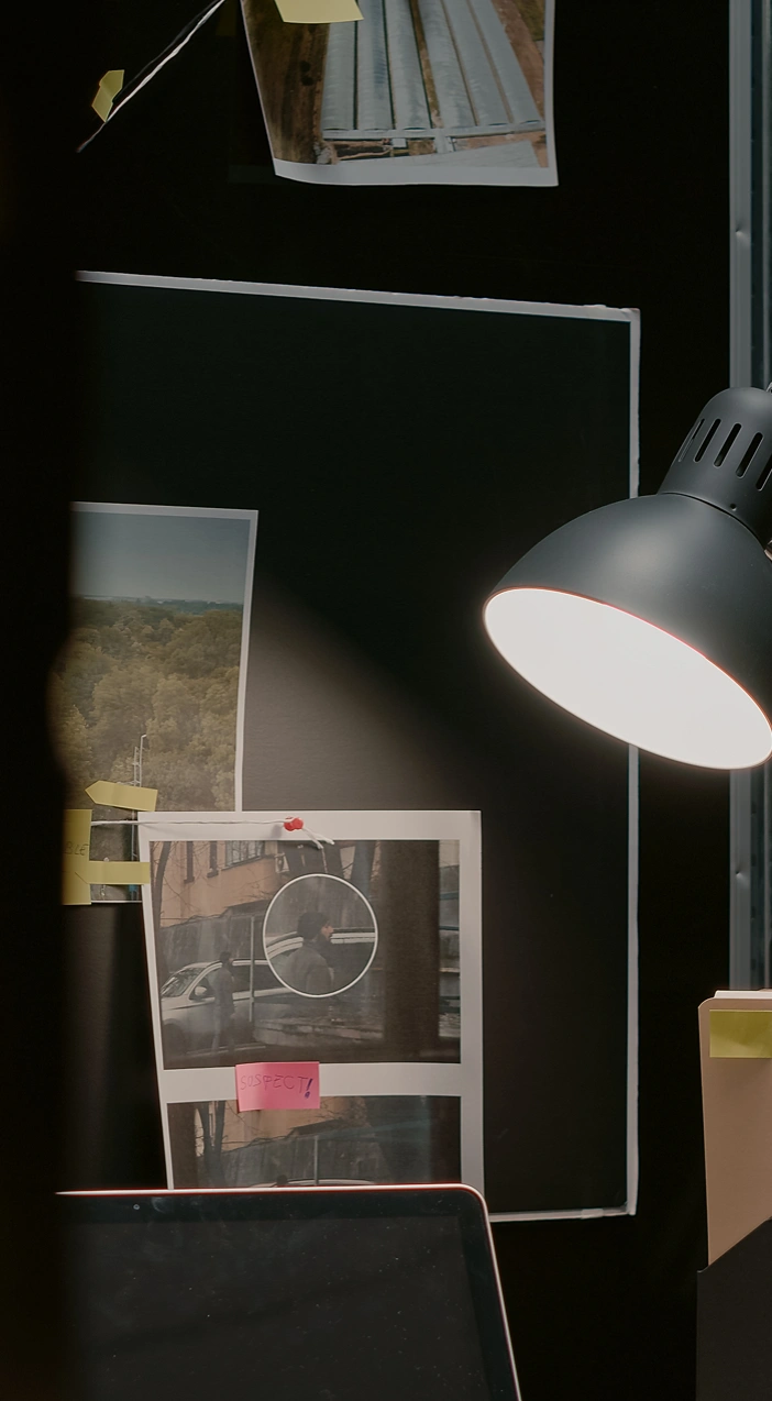 Private Investigator, GPS Tracking | Desk lamp illuminating a surveillance photo marked with a pink 'Suspect' sticky note on a dark investigation board.