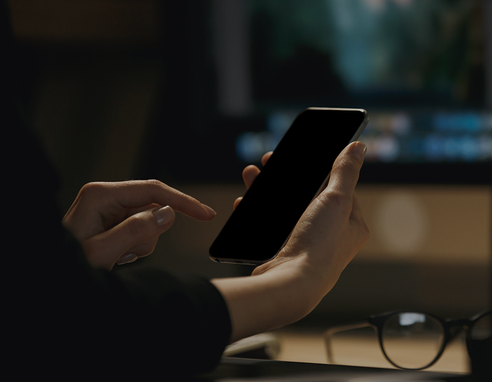 Investigation Services | Close-up of hands holding a smartphone with a blank black screen in a dimly lit room, with glasses resting on the desk.